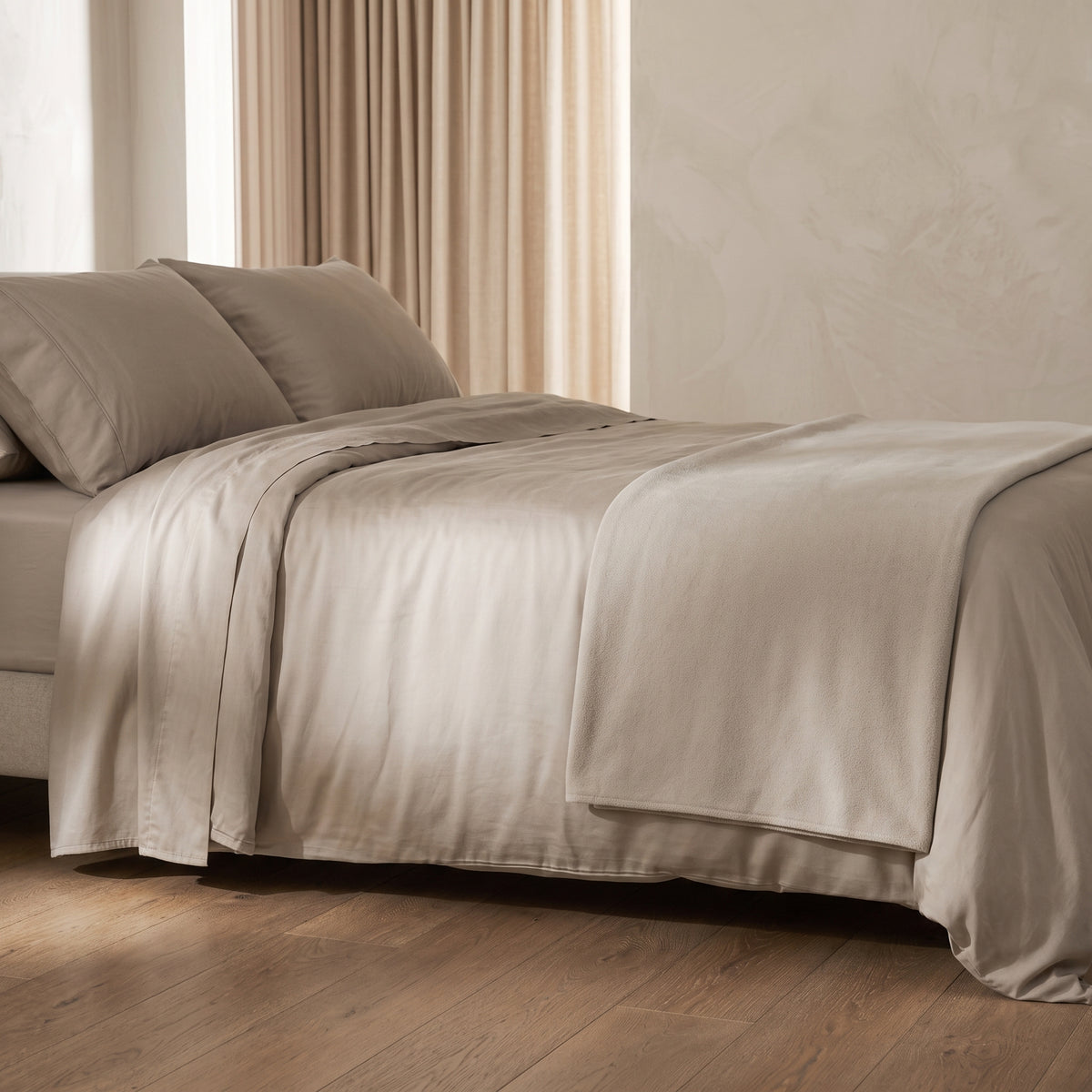 Ein unaufdringlich gestaltetes Bett in einem gedämpft beleuchteten, ruhigen Schlafzimmer, bezogen mit glatter, matt-schimmernder Bambus-Viskose-Bettwäsche in dunklem Schattengrau. Die sanft fallenden Laken und Kissen wirken beruhigend und natürlich im dämmrigen Licht.